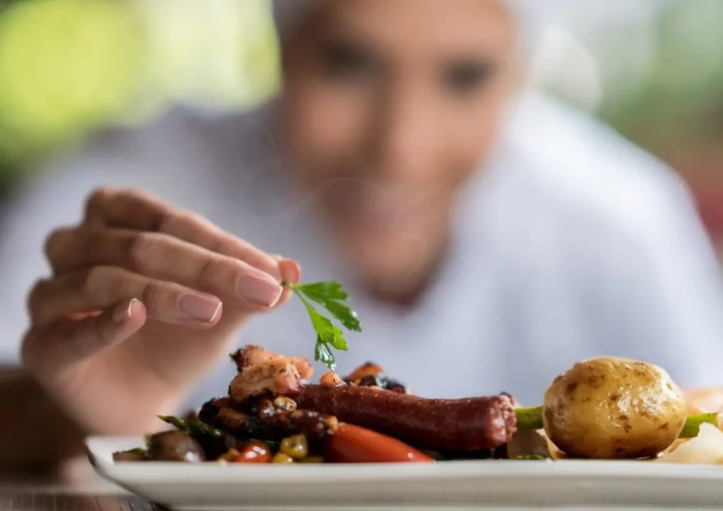 A chef adding a garnish to a plated dish featuring meat, vegetables, and a potato.