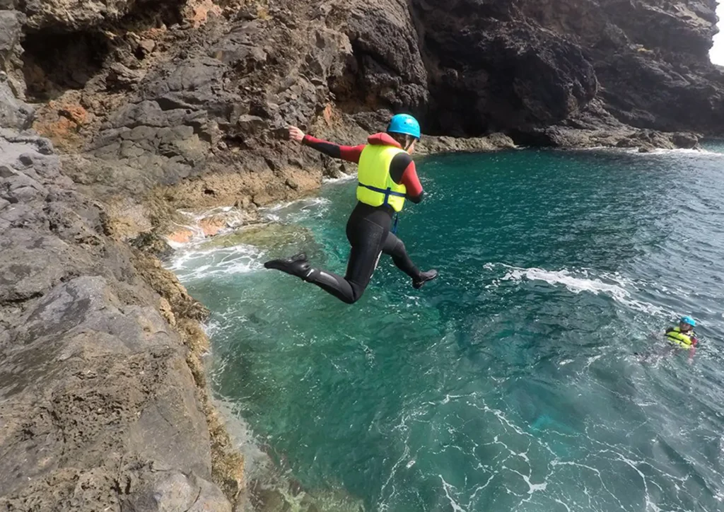 Individual in a wetsuit and helmet jumping off a rocky ledge into the ocean water in Madeira.