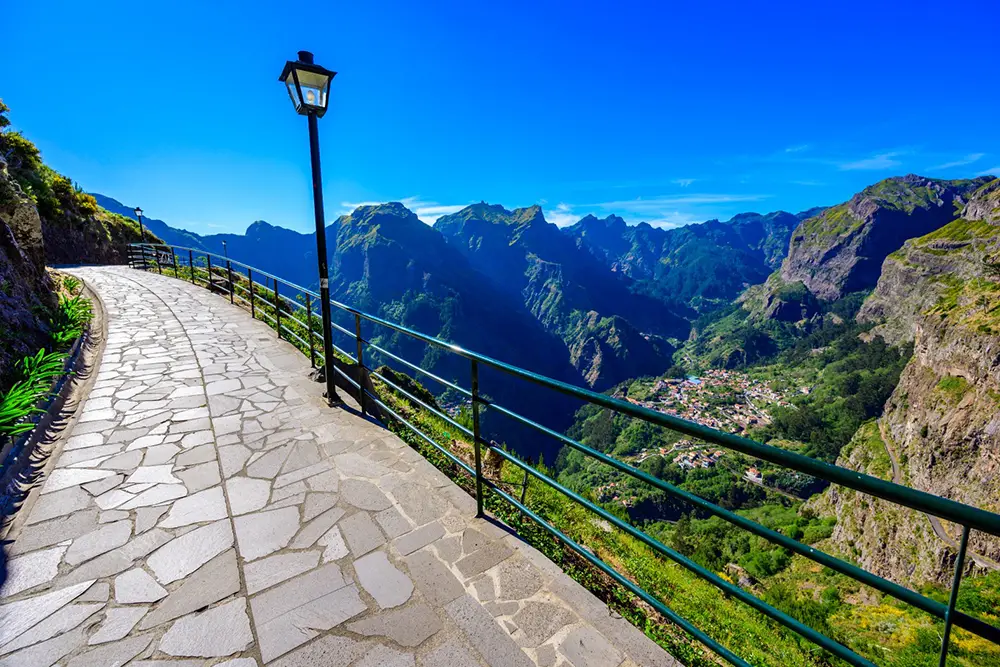 Curved stone pathway with a railing overlooking a mountainous landscape and a village below.