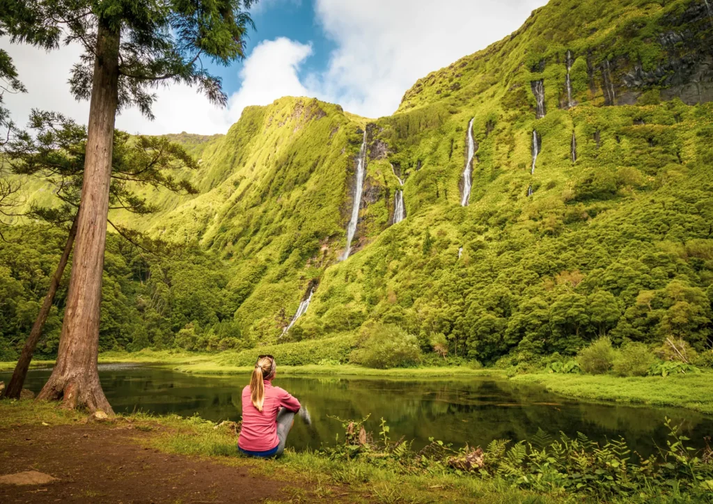 Individual sitting on the shore of a lake, with waterfalls cascading down a green mountain in the background.