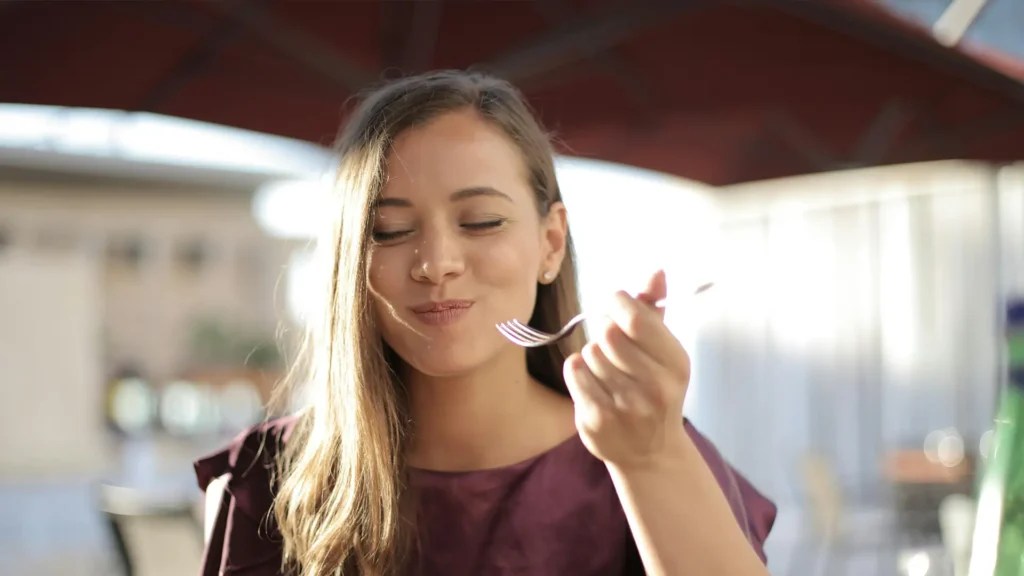 Woman with long hair smiling while eating with a fork outdoors under an umbrella.