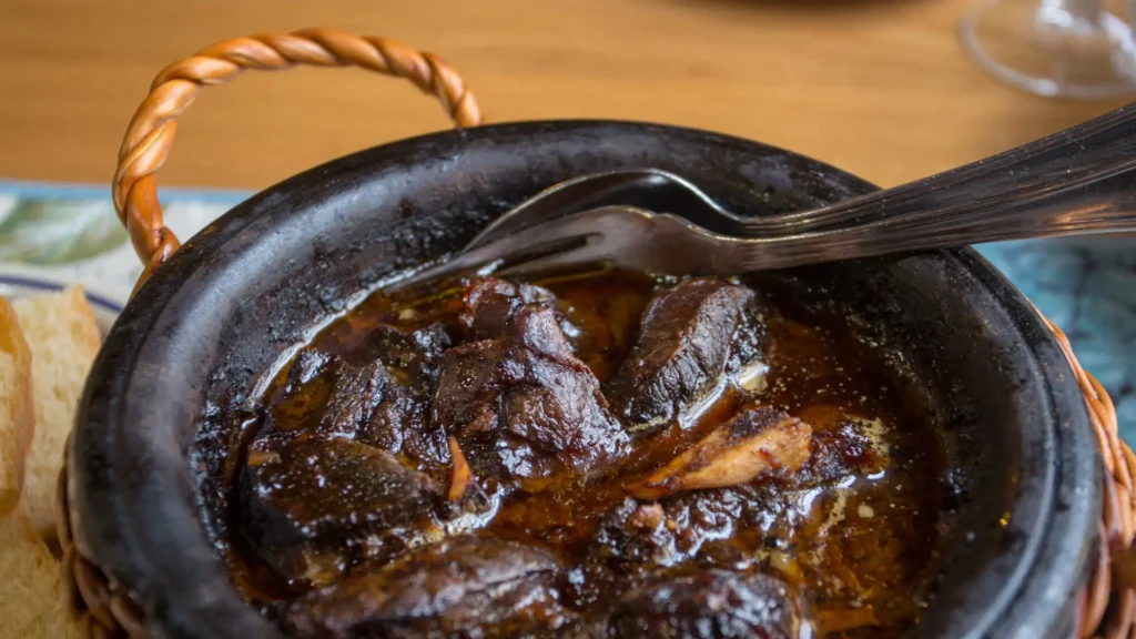 A clay pot filled with cooked meat pieces and sauce, accompanied by a fork resting on the side.