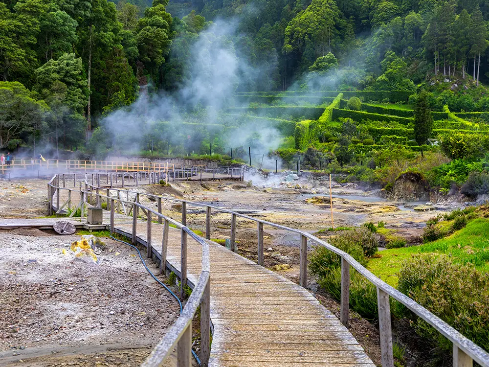 Wooden pathway winding through a geothermal area with steam rising from the ground at Furnas Lake.
