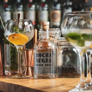Two gin cocktails in glasses with a bottle labeled Rocha Negra and garnishes on a wooden bar.