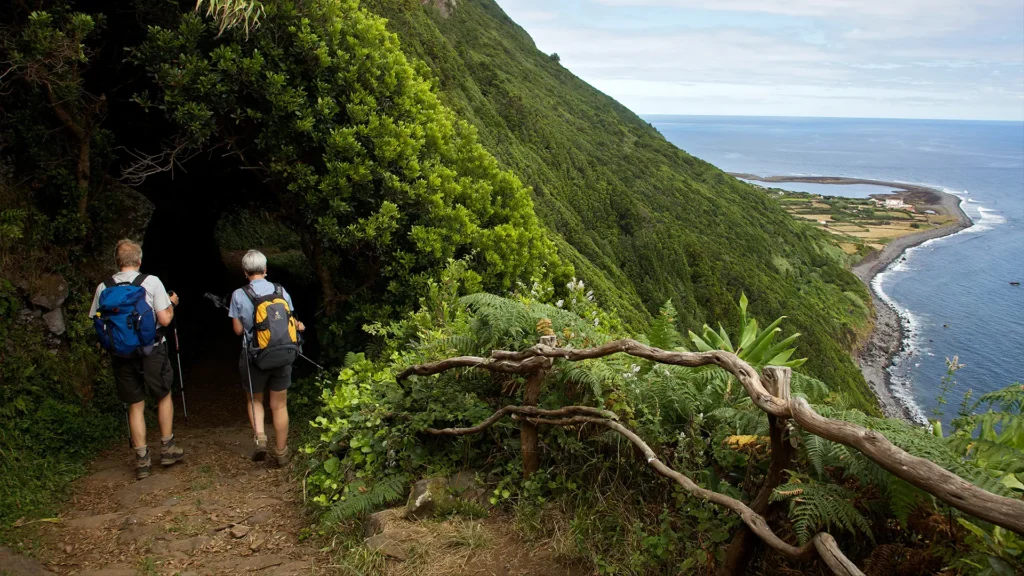 Two hikers walking on a trail with greenery and a coastal view in the background.