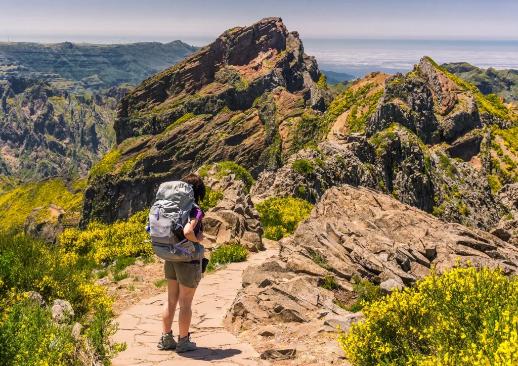 Person with a backpack walking on a rocky trail in a mountainous area with greenery.