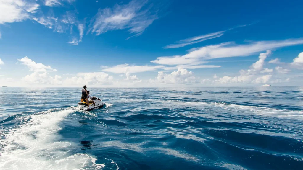 Person riding a jet ski across calm ocean waters under a blue sky with clouds.