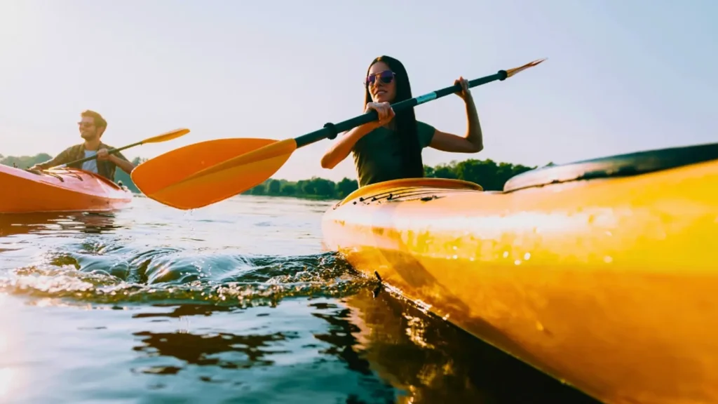 Individual paddling a yellow kayak on calm water with a paddle in hand and sunglasses on.