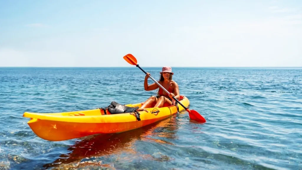 Individual paddling in an orange kayak on calm water under a clear sky.