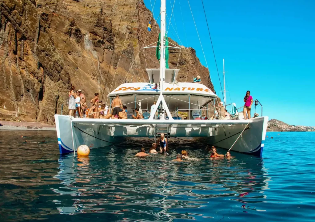A catamaran boat with passengers on board in the water near rocky cliffs in Madeira.