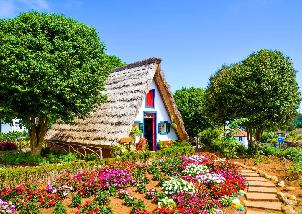 A traditional Madeiran house with a thatched roof and colorful doors in Santana, surrounded by flowers.