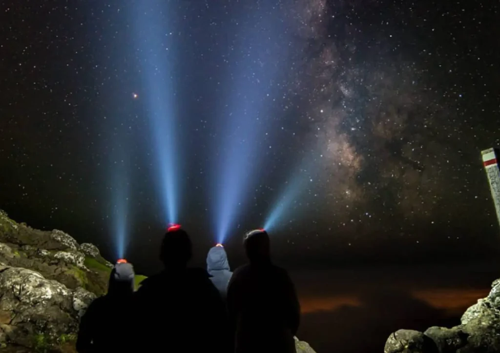 Silhouetted figures with headlamps illuminating the night sky filled with stars and the Milky Way.