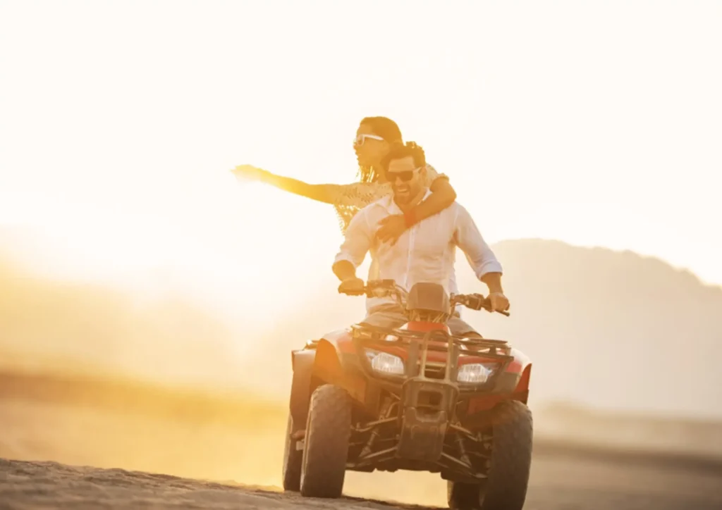 A man and woman riding an all-terrain vehicle during sunset, with the sun setting in the background.