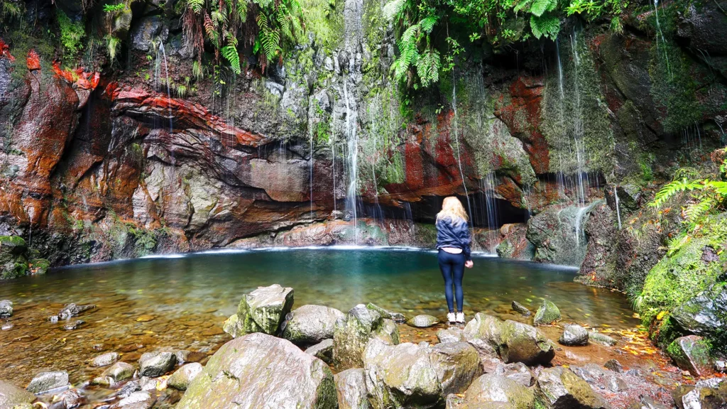 A woman standing by a pool of water in front of a waterfall surrounded by rocks and greenery.