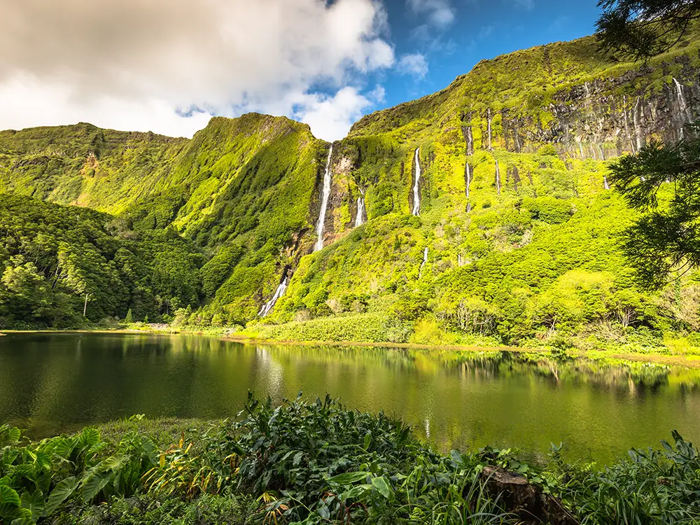 A scenic view of waterfalls cascading down a green mountain into a calm lake surrounded by lush vegetation in Flores Island.