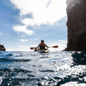Individual kayaking through a narrow coastal cave with rocky cliffs on either side and bright blue water.